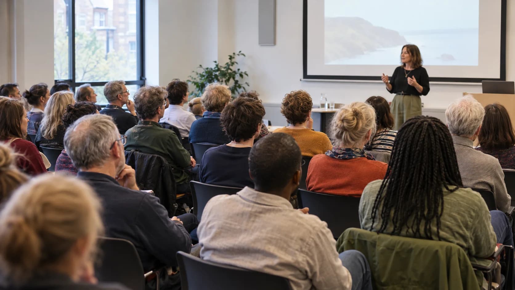 Illustration of an audience of different ages listening to a speaker in a university lecture room
